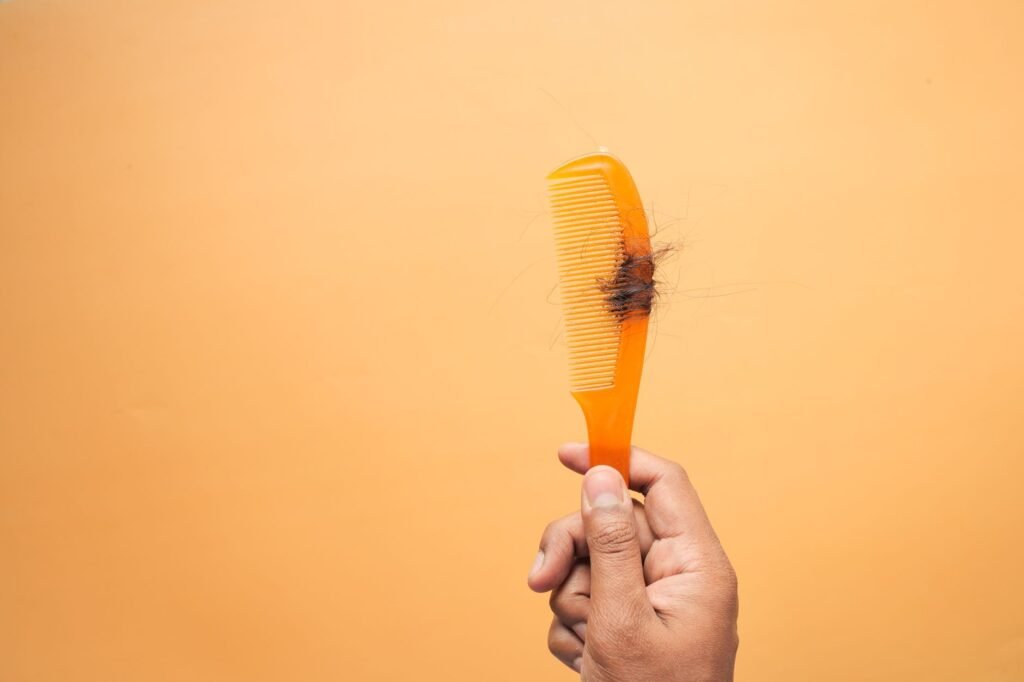 Close Up Of Hand Holding A Comb With Hair Against An Orange Backdrop, Highlighting Hair Loss Concept.