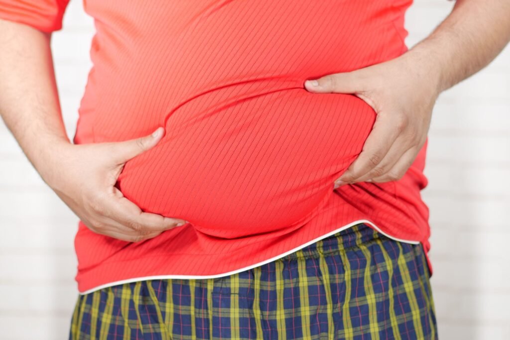 Close Up Image Of A Man Holding His Bloated Belly While Wearing A Red Shirt.